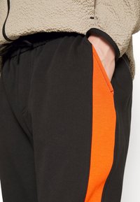 Black joggers with an orange side stripe, made of soft material. Features an elastic waistband and side pocket for functionality.