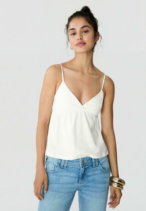 Femme aux cheveux foncés en chignon, portant un haut blanc sans manches à col en V, un jean bleu clair et de larges bracelets dorés au poignet droit.