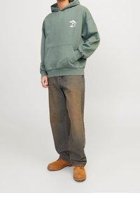 Vihreä huppari, jossa on etutasku ja graafinen kuviointi. Yhdistetty löysiin ruskeisiin farkkuihin ja ruskeisiin mokkanahkasaappaisiin.