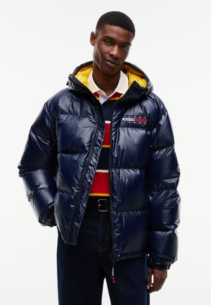 Junger Mann trägt eine glänzende marineblaue gesteppte Jacke mit gelbem Kapuzenfutter, gestreiften Pullover und dunkle Jeans und steht mit den Händen in den Taschen.