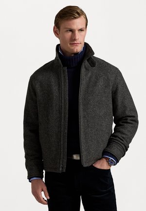 Dark gray wool blend jacket with a stand collar, front zipper, and ribbed cuffs, layered over a navy turtleneck and worn with slim black pants.