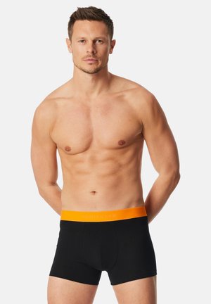 Sorte boksershorts med en skrigende orange talje. Stoffet ser glat og tætsiddende ud, med et logo trykt på taljen. Modellen er overkropsnøgen.
