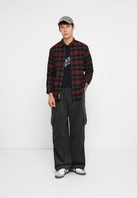 Camisa de franela de cuadros en rojo y negro sobre una camiseta gráfica negra. Pantalones cargo sueltos negros. Gorra gris y zapatillas blancas con acentos negros.