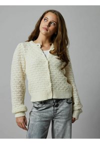 Cardigan bianco lavorato a maglia con un motivo testurizzato, dotato di chiusura a bottoni e polsini a costine. Indossato su una maglietta semplice e abbinato a jeans chiari.