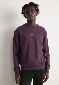 Sudadera morada con cuello redondo, que presenta un pequeño logotipo blanco "BOSS" en el pecho. Combinada con jeans negros, atuendo centrado.