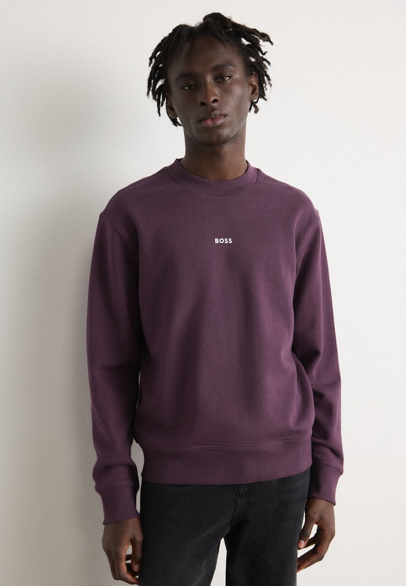 Sudadera morada con cuello redondo, que presenta un pequeño logotipo blanco "BOSS" en el pecho. Combinada con jeans negros, atuendo centrado.