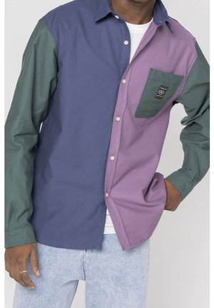 Hombre que lleva una camisa abotonada con bloques de color en morado, verde y azul marino, combinado con vaqueros azul claro.