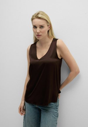 Vrouw met blond haar, gekleed in een donkerbruine mouwloze top met V-hals en lichtblauwe spijkerbroek, staand tegen een effen witte muur.