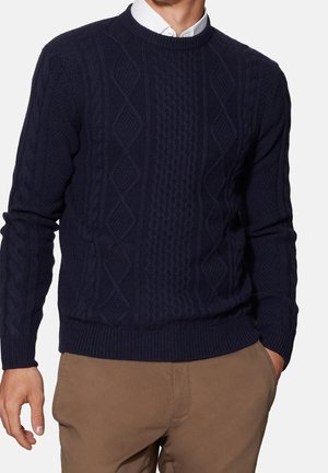 Mann trägt navyblauen Zopfpullover über weißem Hemd mit Kragen und brauner Hose, Hand in der Tasche vor einfarbigem Hintergrund.