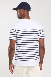 Mand iført en marineblå kasket, en hvid T-shirt med vandrette marineblå striber og lyse bukser, vist fra ryggen mod en ensfarvet baggrund.