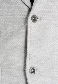 Graue Jacke aus strukturiertem Stoff mit einem sichtbaren Knopf, auf dem die Marke "Tom Tailor" eingeprägt ist, und einem schlichten, klassischen Design.