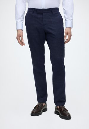 Muž nosi námornícky modré na mieru šité nohavice, bielu košeľu na gombíky a tmavohnedé mokasíny s strapcami, stojí pred jednoliatym svetlým pozadím.