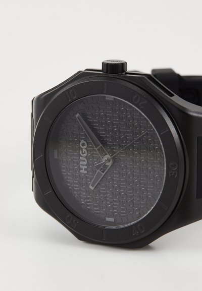 Montre analogique noire avec un cadran noir texturé, logo en relief, aiguilles argentées et un bracelet en résine noire robuste.