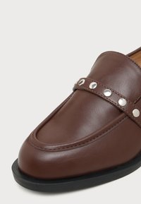 Mocassin en cuir marron avec des clous ronds argentés sur une bride traversant le dessus, semelle noire et coutures visibles le long de la zone des orteils.
