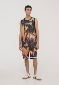 Canottiera in rete con stampa di un tramonto e palme, abbinata a pantaloni corti coordinati. Colori nero e arancione con accenti bianchi. Sneakers bianche.