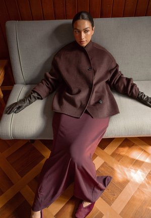 Burgundy wool coat with a high collar, paired with a matching flowing dress and dark leather gloves, seated on a gray sofa.