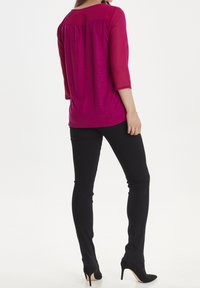 Top fucsia con maniche trasparenti a 3/4, vestibilità rilassata e pieghe posteriori, abbinato a pantaloni neri attillati e tacchi alti neri.
