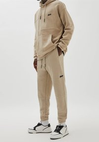 Sweat à capuche beige avec cordon de serrage ajustable, coupe oversize et poche avant ; pantalon de jogging beige assorti avec panneaux texturés ; porté avec des baskets noires et blanches.