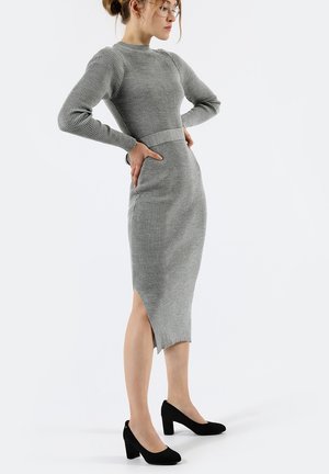 Abito maglione grigio a coste con maniche lunghe, vita aderente e spacco laterale. Abbinato a scarpe con tacco blocco nere. Design semplice e raffinato.