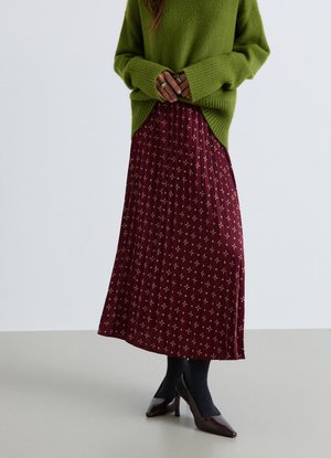 Donna che indossa un maglione verde oversize, una gonna plissettata color granata con un piccolo motivo floreale, calzini neri e tacchi marrone scuro.
