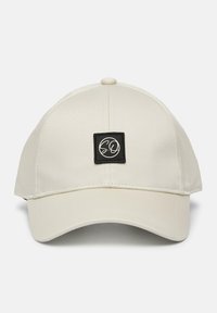 Lichtbeige baseballcap met een gebogen rand en een vierkante zwarte patch met een wit logo. Glad stof met stikselaccenten.