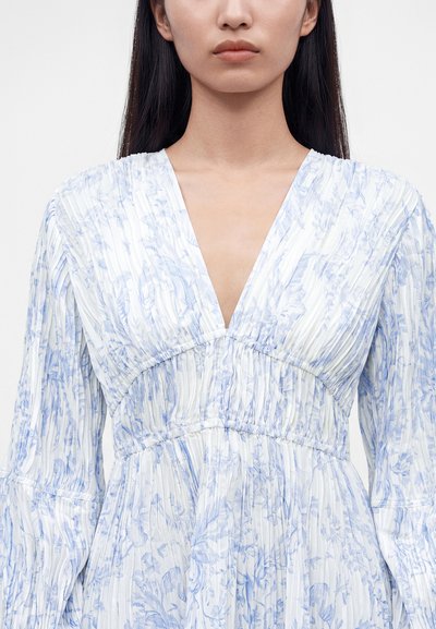 Femme aux longs cheveux foncés portant une robe blanche à motif floral bleu et un décolleté en V profond sur un fond clair uni.