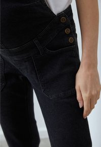 Svarta denimoverall med en figurnära design, tre bronsknappar på sidan och framfickor med en mjuk textur.