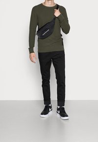 Maglione verde oliva con texture e dettagli a righe, jeans neri slim-fit e sneakers nere con accenti bianchi. Borsa a tracolla nera con logo.