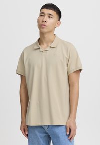 Beige pólóing, pamutból készült, hajtogatott gallérral, rövid ujjú, a mellkasán kis logó részlettel, kék farmernadrággal viselve.