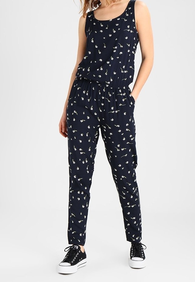 Femme portant une combinaison bleu marine sans manches avec un petit imprimé floral blanc et des baskets noires, debout les mains dans les poches.