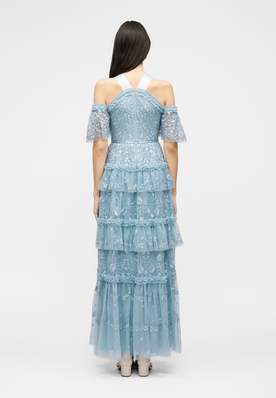 Γυναίκα με μακριά σκούρα μαλλιά, φορώντας ένα μακρύ φούστα με τούλι σε ανοιχτό μπλε χρώμα και μανίκια με ακάλυπτους ώμους, στέκεται με την πλάτη στην κάμερα.