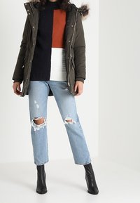 Parka vert olive avec bordure en fourrure sur la capuche, pull en tricot à motif coloré noir et orange, jeans en denim clair déchirés, et bottines noires.
