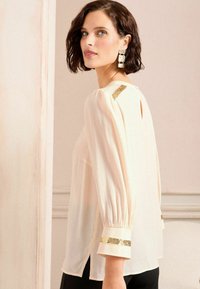 Blusa color crema chiara con maniche arricciate, strisce dorate decorative ai polsini e alla parte posteriore del collo, e un'apertura a chiave sul retro.