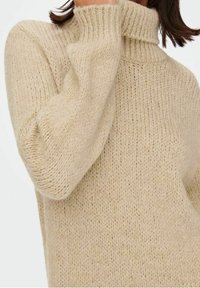 Femme portant un pull en tricot côtelé beige, la main couvrant partiellement sa bouche.
