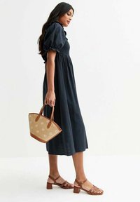 Vestido azul marino de longitud media con mangas abullonadas y cuerpo fruncido; sandalias marrones con tiras; bolso de mimbre con acentos florales.