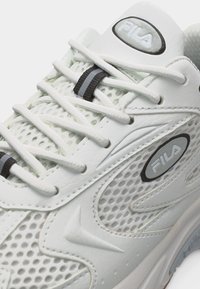 Λευκό αθλητικό παπούτσι FILA με λεπτομέρειες από δίχτυ και δέρμα, λευκά κορδόνια με μαύρες και γκρι ρίγες στους κλειδαριές κορδονιών, κοντινή όψη στα λογότυπα στο πλάι και στη γλώσσα.