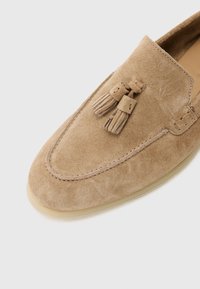Beige ruskinn loafers med dekorative dusker, rund tå og synlig søm. Gjennomsiktig gummisåle. Glatt tekstur.