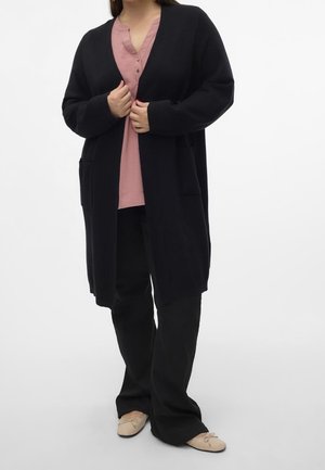 Femme portant un long cardigan noir ouvert sur un haut rose à boutons, un pantalon noir et des ballerines beiges, debout avec les mains tenant le cardigan.