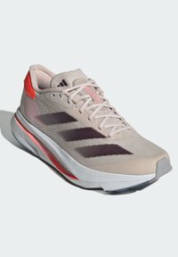 Světle béžová běžecká obuv Adidas s tmavými třemi pruhy, růžovými akcenty, bílou podrážkou a tkaničkami, zobrazena nakloněná doprava na bílém pozadí.