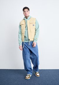 Beige Utility-Weste über einem mintgrünen Hoodie, kombiniert mit lockeren blauen Jeans und mehrfarbigen Turnschuhen. Einfaches Design mit mehreren Taschen.