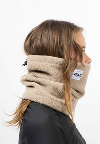 Tour de cou en polaire beige avec une surface texturée et un cordon de serrage ajustable. Présente une patch logo sur le côté, porté par-dessus des vêtements sombres.