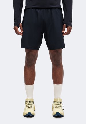 Homme portant un short de sport noir, des chaussettes blanches et des chaussures de course beige, debout sur un fond clair.