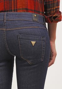 Dunkelblaue Denim-Jeans mit einem leicht taillierten Design, das orangefarbige Nähte und ein dreieckiges goldenes Logo auf der Gesäßtasche aufweist.