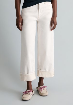 Osoba nosząca kremowe, szerokie spodnie 7/8 z podwiniętymi mankietami oraz beżowe wsuwane buty z fioletowymi sznurowadłami, stojąca na tle w kolorze szarym.