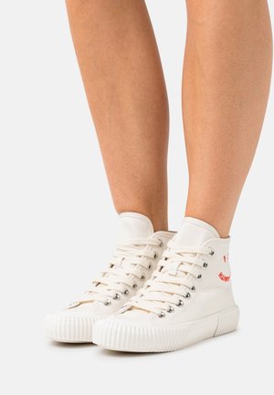 Nohy v béžových vysokých plátěných teniskách s vroubkovanými podrážkami a bílými tkaničkami, stojící na světlešedém povrchu.