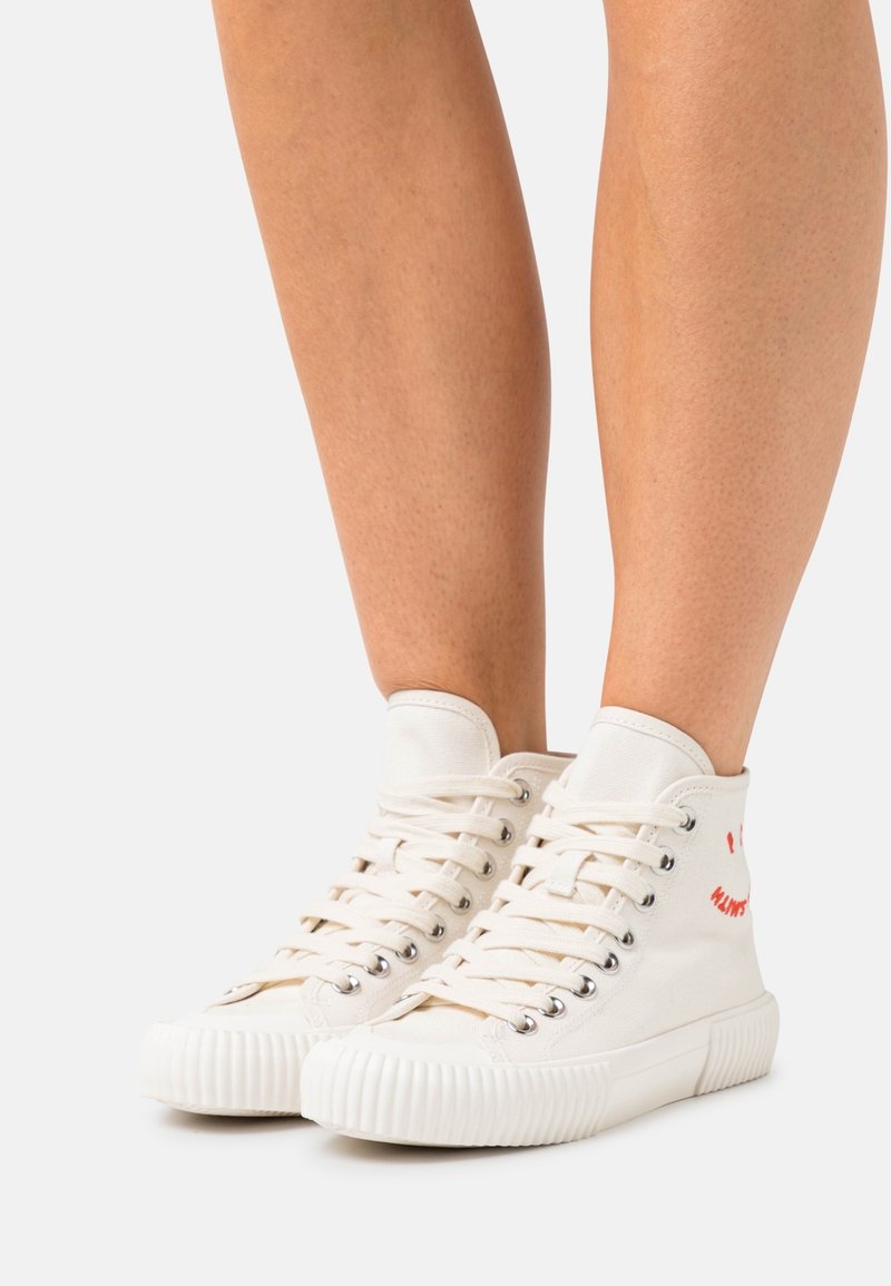 Jambes portant des baskets montantes en toile blanc cassé avec des semelles côtelées et des lacets blancs, se tenant sur une surface gris clair.