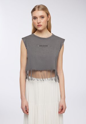 Giovane donna con lunghi capelli biondi, che indossa un top corto grigio senza maniche con frange e una gonna plissettata bianca, in piedi davanti a uno sfondo bianco.