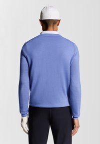 Maglione lavorato a maglia blu chiaro con maniche lunghe, caratterizzato da polsini e orlo a costine, indossato con pantaloni scuri e un cappellino bianco. Texture morbida.