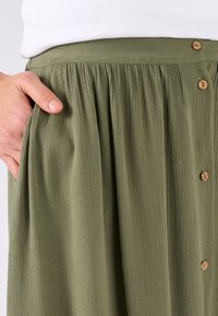 Falda plisada de color verde oliva, hecha de tejido ligero con detalles de botones de madera y bolsillos laterales. La superficie texturizada añade interés visual.