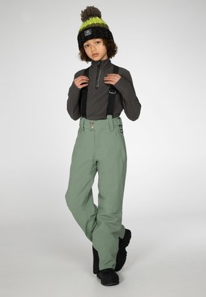 Pantaloni da sci verde menta con bretelle nere regolabili, abbinati a una maglietta a maniche lunghe grigio scuro e un cappello di maglia nero con un pompon verde.